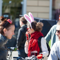 People are gathered outdoors in bright sunlight, some standing next to bicycles. The focus is on a person wearing white rabbit ears and a red scarf, reminiscent of Kritiskā masa 2012. Around this individual, others engage in conversation—one donned sunglasses and another a red helmet—with a blurred building in the background.