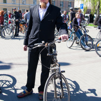 A man with a beard and mustache stands outdoors, holding a bicycle during the 2012 Kritiskā masa event. He is wearing a black suit with a white dress shirt and colorful shoes. Several other people with bicycles are in the background, with buildings and trees visible on this sunny day.