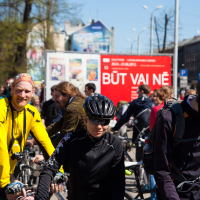 A group of cyclists gathered in an urban area, mostly wearing helmets and sports attire. One cyclist in a yellow jacket stands out, while others blend in with darker clothing. Behind them is a large red billboard with white text and surrounding people, buildings, and trees in the background from Kritiskā masa 2012.