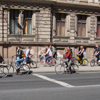A group of people ride bicycles on a city street next to an old, weathered building with tall, narrow windows. Cyclists of various ages and genders move in the same direction, reminiscent of the Kritiskā masa events from 2012. Sunlight casts shadows, emphasizing the clear weather.