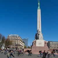 A tall monument with a statue holding three stars stands in an urban square under a clear blue sky. Cyclists and pedestrians, reminiscent of the 2012 Kritiskā masa movement, are actively moving around the square. Historic buildings with ornate architecture are visible in the background, along with a modern high-rise.