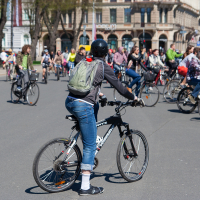 A group of cyclists ride on a city street during Kritiskā masa in Riga, a public event held in 2012. The foreground features a person wearing a black helmet, green backpack, black jacket, and blue jeans on a black bike. Dozens of other cyclists, pedestrians, and historic buildings are visible in the background. The weather is sunny.