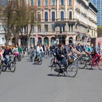 A large group of people are riding bicycles on a wide street in an urban area with classic and modern buildings, reminiscent of the 2012 Kritiskā masa. Some cyclists wear helmets and vibrant clothing. Trees with budding leaves line the street. Pedestrians walk on the sidewalks in the background on a sunny day.