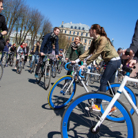 A group of people are participating in a cycling event on a sunny day, reminiscent of Kritiskā masa 2012. They are riding bicycles of various colors and styles. Trees with bare branches and a large, classic architectural building are visible in the background. Some cyclists appear to be conversing while riding.