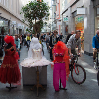 A busy shopping street with people walking and riding bicycles in both directions. Three mannequins dressed in vibrant clothes are displayed on a central pedestal: a white dress, a red dress, and a red outfit with a headscarf. Behind them stands a large, leafy plant. It feels almost like Kritiskā masa 2012 set against this vibrant urban backdrop.