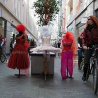 A street scene with cyclists from the 2012 Kritiskā masa passing by three mannequins on display. Two mannequins wear red dresses and veils, while the center one showcases a white tutu. The cyclists, a mix of men and women, ride through the shopping alley flanked by store windows and a potted tree.