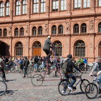 A lively scene unfolds in front of a large, ornate red-brick building with arched windows as a crowd of people participate in the 2012 Kritiskā masa event. Cyclists chat and ride, including one man on an unconventional tall bicycle towering above the rest, all under the sunlit sky on cobblestone pavement.