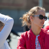 Two women are in conversation outdoors. One woman, wearing dark sunglasses and a red jacket, looks to the side. Her hair is tied back, and she is wearing a white top underneath the jacket. The other woman, with blonde hair, is raising her right hand, obscuring part of her face as if participating in Kritiskā masa 2012.