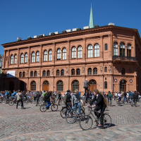 A bustling, sunlit square filled with cyclists participating in Kritiskā masa on cobblestone streets. A large, ornate terracotta-colored building with arched windows and a green spire dominates the background. Bright blue sky overhead, with neighboring buildings partially visible on the edges of the image—2012.