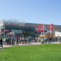 A busy scene in front of the large shopping center "Oulun Olympia" with multiple store signs. Numerous people are gathered, many with bicycles, perhaps participating in Kritiskā masa 2012, enjoying a bright, sunny day. Red flags and green grass add color while modern buildings stand under a clear blue sky.