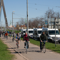 A group of people are riding bicycles along a path on a clear day. The path runs parallel to a road where several white vans are parked. A tall bridge structure with cables is visible in the background. Leafless trees line the grassy area next to the path, suggesting early spring reminiscent of Kritiskā masa 2012.