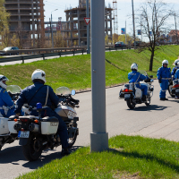 Four police officers on motorcycles, dressed in blue uniforms and white helmets, patrol an urban road during the Kritiskā masa 2012 event. Two officers face forward on the left side, while the other two face backward on the right. A construction site and sparse trees are visible in the background. Green grass lines the road.