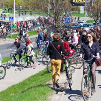A large group of people participating in the 2012 Kritiskā masa bike ride event on a sunny day. Men, women, and children walk with and ride bicycles on a sidewalk and grass area along a busy road. Some participants are smiling, and there are trees and buildings visible in the background.