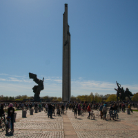 A large outdoor plaza bathed in sunlight features two prominent statues on either side and a tall central monument. Numerous people with bicycles, reminiscent of Kritiskā masa 2012, are gathered, enjoying the day under a clear blue sky. Trees in the background are slightly autumnal, offering a splash of green and yellow hues.