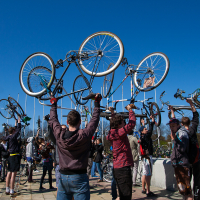 A group of people are gathered outdoors on a sunny day, raising their bicycles above their heads in a celebratory manner. The sky is clear and blue, and people are dressed in casual clothing. Some individuals are seen smiling and interacting with each other, reminiscent of the Kritiskā masa event in 2012.