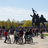 A large group of people gather on a cobblestone square with bicycles, reminiscent of the 2012 Kritiskā masa event. In the background, there's a tall, abstract sculpture of several figures. Trees with light green foliage mark the horizon under a clear blue sky. The relaxed crowd is engaged in conversations and activities.