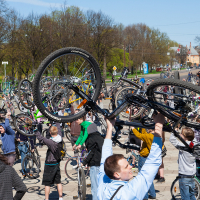 A large group of people gathered outdoors in a park on a sunny day, many holding up bicycles over their heads. The crowd is diverse, and trees in the background are budding, suggesting springtime. This 2012 event appears to be Kritiskā masa, celebrating or demonstrating the joys of cycling.