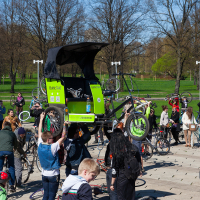 A lively scene of a large group of people gathering outdoors in a park for the 2012 Kritiskā masa event. Some are holding up their bicycles, and a green pedicab with a black canopy is prominently displayed in the center. The weather appears sunny, and the background features leafless trees and green grass.