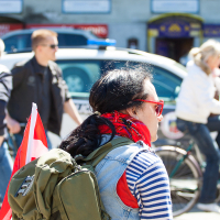 A person with a black ponytail, wearing red sunglasses, a striped shirt, and a green backpack with a red-and-white flag attached, is seen from behind during the 2012 Kritiskā masa. They are part of a group of cyclists. Several people on bicycles and a police car are in the background on a sunny day.