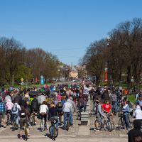 A large crowd of cyclists and pedestrians, viewed from behind, participate in the 2012 Kritiskā masa event on a sunny day. The group is moving along a broad pathway lined with leafless trees. Participants wear casual attire, and some carry backpacks. Blue skies and distant buildings are visible in the background.