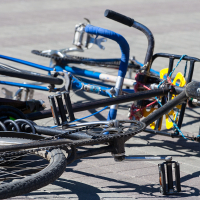 Two bicycles lie on the ground on a paved surface during Kritiskā masa 2012. One has a blue frame with black handlebar tape and no visible front wheel, while the other has a black frame with the pedal and gear area prominently showing. A black backpack is partially visible near the bicycles.