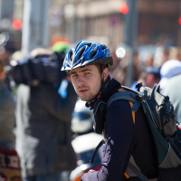 A cyclist wearing a blue helmet, black long-sleeve shirt, and a backpack pauses amidst a busy city street during Kritiskā masa 2012. He looks ahead while people around him, some also on bikes, engage in various activities. The background is blurred, showing a mix of pedestrians and urban elements.