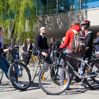 A group of people with bicycles gather outside under a sunny sky. The scene, reminiscent of Kritiskā masa 2012, includes both men and women dressed in casual attire, conversing animatedly. A modern building with large windows and a tree with green leaves form the background, creating a lively and relaxed atmosphere.