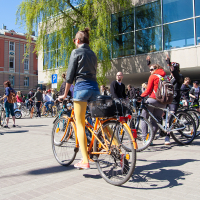 A group of people with bicycles gather in a sunny urban plaza near a modern glass building. One person with orange tights and a denim jacket stands prominently in the foreground holding a yellow bicycle. The crowd, reminiscent of the 2012 Kritiskā masa event, appears relaxed, some standing and talking, others seated on their bikes.