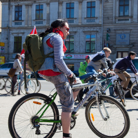 A cyclist stands beside their bicycle on a city street, preparing for a ride. They are wearing a striped long-sleeve shirt, camouflage shorts, and red sneakers with white stars. A green backpack and a red flag are attached to the bike. Other cyclists and historic buildings are visible in the background, reminiscent of Kritiskā masa 2012.