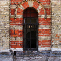 An arched window set in a weathered brick and stone wall of Daugavgrīva, protected by rusty iron bars, revealing a dark, obscured interior behind shattered glass panes.