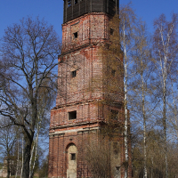 An old, weathered red brick Daugavgrīva tower stands tall against a clear blue sky, flanked by slender birch trees with early spring foliage.