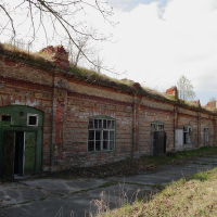 An abandoned red-brick building in Daugavgrīva, with deteriorating walls and missing doors under a partly cloudy sky, indicative of neglect and the passage of time.