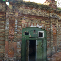 An old, weathered brick structure in Daugavgrīva with a damaged green door ajar, revealing a dark interior, beneath a clear, blue sky.