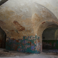 An old, abandoned interior space in Daugavgrīva with faded and peeling turquoise paint, featuring a series of brick and plaster archways leading into darkness, hinting at a history long
