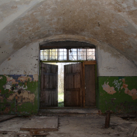 An abandoned room in Daugavgrīva with peeling green walls, a vaulted ceiling, and a deteriorating wooden door under a barred window, conveying a sense of decay and forgotten history