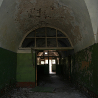 A dilapidated hallway with peeling paint and debris in Daugavgrīva, featuring an arched ceiling and a fractured wooden floor, suggesting neglect and the passage of time within an abandoned