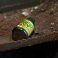 An old, dusty bottle labeled "tinctura valerianae" lies on a rusty metal surface in Daugavgrīva, surrounded by debris, evoking a sense of neglect and