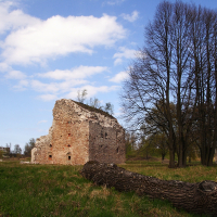 A weathered brick ruin stands in a lush field beside a fallen tree, with bare trees and a cloud-dappled blue sky overhead in Daugavgrīva, evoking a sense of