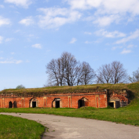 An old red brick fortification with arched entrances is partially overgrown with grass, set against a backdrop of a clear blue sky and a sparse tree line near Daugavgrīva.