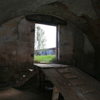 A dilapidated wooden ramp in Daugavgrīva leads to an arched doorway framing a view of an overgrown yard and abandoned buildings, evoking a post-apocalyptic atmosphere.