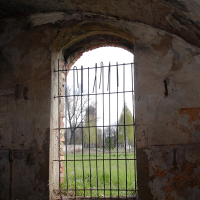 An arched, barred window in a weathered brick wall at Daugavgrīva offers a view of a green park, juxtaposing confinement with the freedom of nature outside.