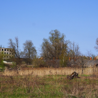 Abandoned buildings in Daugavgrīva overgrown with vegetation under a clear blue sky, showing nature's reclamation of a once-industrial area.