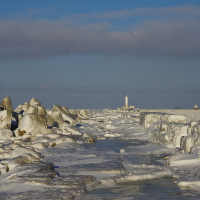 A winter scene in Daugavgrīva featuring ice-covered rocks and a lighthouse.
