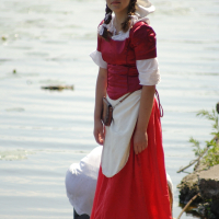A young woman stands by a lake, dressed in a red historical costume with a white apron and headscarf. She has braided hair and wears lace-up shoes. The calm waters of the Dunamunde shoreline provide a serene backdrop, while another person kneels near the water, mostly out of view.