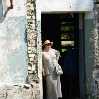 A person wearing a beige outfit and a straw hat stands in the doorway of a partially dilapidated Dunamunde 2010 building with peeling paint and exposed bricks. They have a white bag across their shoulder and are looking out into the sunlight. The area around the building appears grassy and untended.