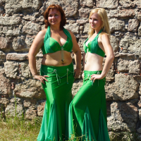 Two women stand in front of a stone wall, wearing matching green belly dance costumes, reminiscent of Dunamunde 2010. The woman on the left has short red hair, and the woman on the right has long blonde hair. Both are smiling and posing with one hand on their hip. Grass is visible at the bottom of the image.