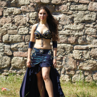 A woman with long curly hair stands in front of a stone wall, reminiscent of Dunamunde 2010. She is wearing a dark blue belly dance costume adorned with gold embellishments. The outfit includes a top, arm bands, and a skirt with a high slit, revealing part of her leg. Grass is visible at her feet.