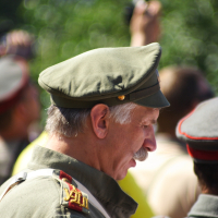 A man with a gray mustache wearing a vintage military uniform and a matching cap stands outdoors with his head slightly bowed at the Dunamunde 2010 event. The uniform features red and gold shoulder epaulettes. Blurred figures in similar attire are visible in the background, suggesting a group or gathering.