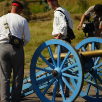 Several people in historical military attire stand around a bright blue cannon with large wheels in an outdoor setting. Some are adjusting the cannon, while another person in modern clothing appears to be filming the scene, likely capturing the 2010 Dunamunde reenactment. Dry grass and stacked hay are visible in the background.