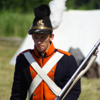 A man dressed as a historical soldier stands outdoors at Dunamunde 2010, holding a rifle with a bayonet. He's wearing a navy blue and orange uniform with white crossbelts, a tall shako hat adorned with a black and white plume, and brass insignia. The background features blurred greenery and a white tent.